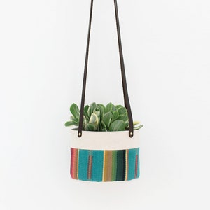 Könnte beinhalten: Ein Hängepflanztopf mit einer weißen Canvas-Basis und einem bunten gewebten Muster. Der Topf ist mit einer grünen Sukkulente gefüllt und hängt an einem braunen Lederriemen.