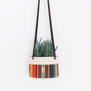 May include: A hanging planter with a woven fabric design in shades of orange, brown, and blue. The planter is filled with a green succulent plant and hangs from a brown leather strap.