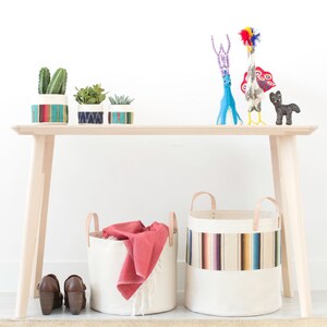 May include: A light wood bench with two canvas storage baskets with colorful stripes. The baskets are filled with a red blanket and a variety of items. There are three small potted plants on the bench, and a blue deer and a colorful bird figurine on the bench.