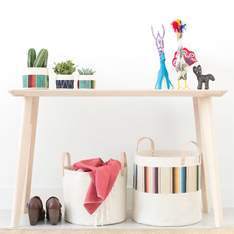 May include: A light wood bench with two canvas storage baskets with colorful stripes. The baskets are filled with a red blanket and a variety of items. There are three small potted plants on the bench, and a blue deer and a colorful bird figurine on the bench.