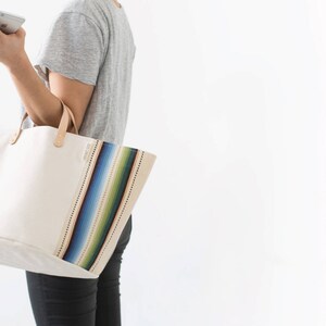 May include: A white canvas tote bag with colorful striped accents. The bag has a brown leather handle and is large enough to carry everyday essentials.
