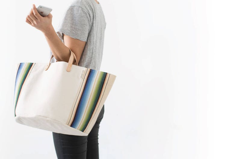 May include: A white canvas tote bag with colorful striped accents. The bag has a brown leather handle and is large enough to carry everyday essentials.