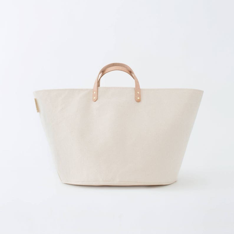 May include: A large, off-white canvas tote bag with brown leather handles and brass accents.