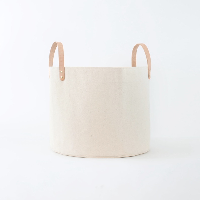 May include: A large, off-white canvas storage basket with two brown leather handles. The basket has a rounded bottom and is perfect for organizing and storing items.