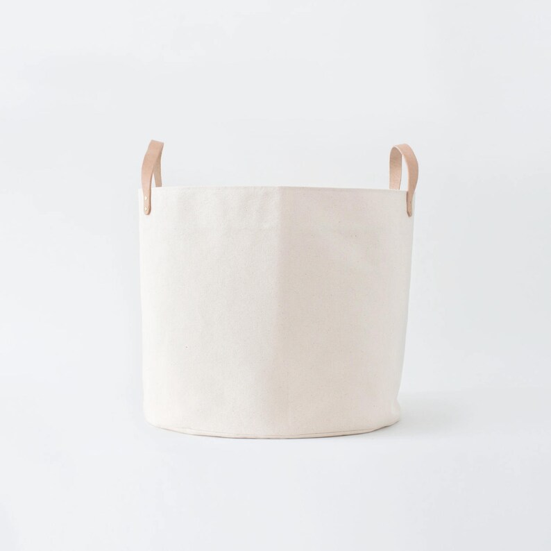 May include: A white canvas storage basket with two leather handles. The basket is empty and sitting on a white background.
