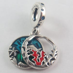May include: A silver charm with a round, openwork design and a smaller, hinged disc. The disc features a blue and green enamel scene, while the openwork side has red accents. The charm has a loop with blue accents.