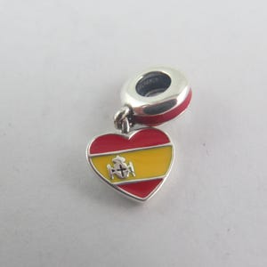 May include: A silver charm bead featuring a heart-shaped pendant with red and yellow enamel stripes. The heart displays a small silver emblem in the centre. The charm has a circular silver bail with a red enamel band around the outer edge.