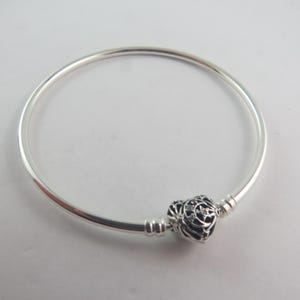 Könnte beinhalten: Ein silbernes Armreif-Armband mit einem herzförmigen Anhänger. Der Anhänger ist schwarz mit einem filigranen Design und kleinen, klaren Steinen. Das Armband hat ein schlichtes, rundes Design, geeignet für den Alltag.