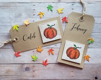 May include: Two brown paper place cards with autumn-themed decorations. The top card has the name "Johnny" written in black cursive. The bottom card has a drawing of an orange pumpkin. The card has the name "Caleb" written in black cursive. Both cards are decorated with small orange, yellow, and green paper leaves.