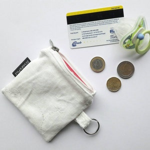 May include: White zippered pouch with a red zipper and a white cord loop. The pouch has the text "TESSIESTYLE" printed on the side. There are four coins and a pacifier on a white surface.