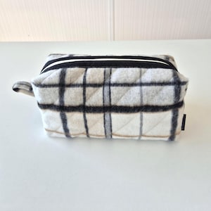 May include: A small, rectangular toiletry bag with a quilted plaid pattern in shades of white, gray, and black. The bag has a black zipper with a white stripe and a small loop handle. The bag is sitting on a white surface.