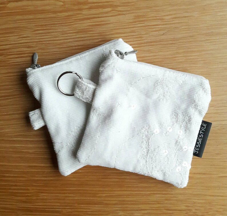 May include: Two white zippered pouches with a key ring. One pouch is plain white corduroy and the other has white embroidery and sequins. The pouch with the embroidery has a black label that says "JESSIESTYLE".