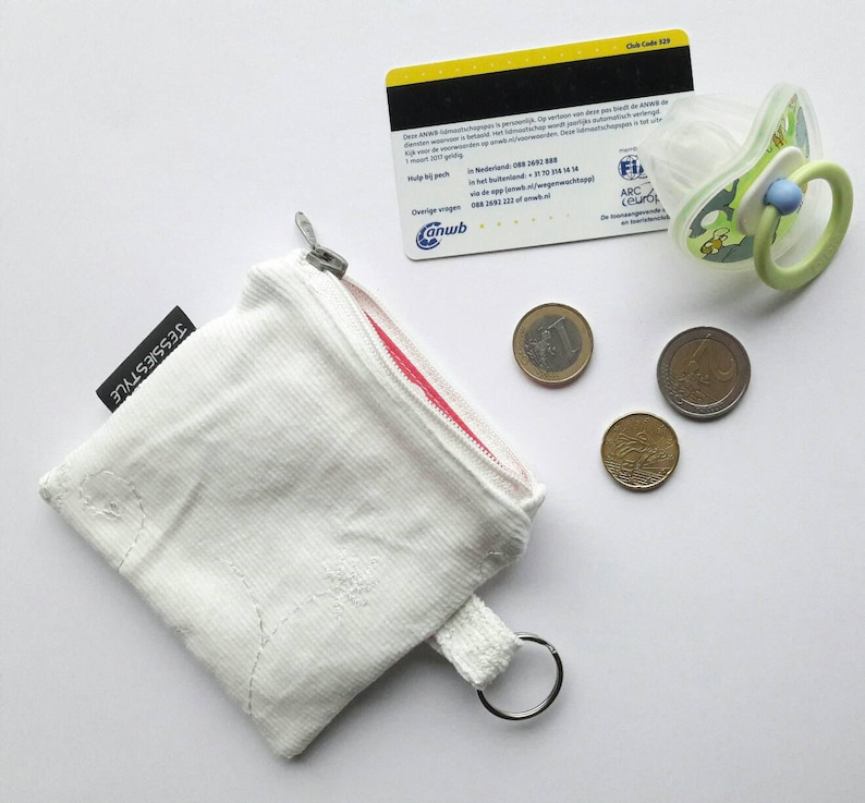 May include: White zippered pouch with a red zipper and a white cord loop. The pouch has the text "TESSIESTYLE" printed on the side. There are four coins and a pacifier on a white surface.