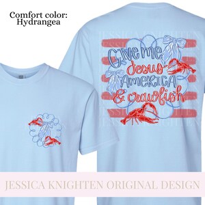 Puede incluir: Camisetas de color azul claro con un dise&ntilde;o que incluye el texto "Give me Jesus America & crawfish". El dise&ntilde;o tambi&eacute;n presenta cangrejos rojos y lazos blancos. La camiseta es de color confort Hydrangea.