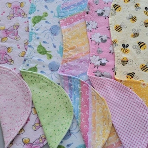 May include: A collection of colorful, patterned baby bibs. Designs include pink elephants, green turtles, blue jellyfish, sheep, bees, and daisies. Each bib has a scalloped edge and is made of soft fabric.