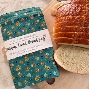 May include: A teal beeswax-lined bread bag with a floral pattern, next to a sliced loaf of bread on a wooden cutting board. The bag has text that reads "Four Acre Homestead Beeswax Lined Bread bag". A bread knife is also visible.