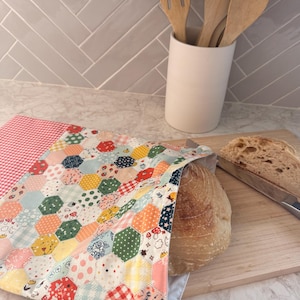 May include: A fabric bread bag featuring a colorful patchwork of hexagonal patterns in shades of red, yellow, green, and blue, with a red and white gingham top section and a white drawstring closure, containing a round loaf of crusty sourdough bread.