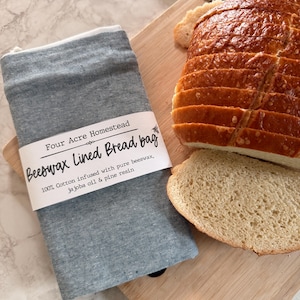 May include: A blue and white striped beeswax-lined bread bag with a loaf of sliced bread and a bread knife on a wooden cutting board. The bag has a label that reads "Four Acre Homestead Beeswax Lined Bread bag".