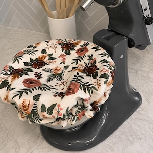 May include: A round, elasticized fabric cover featuring a floral pattern with dark red, peach, and green botanical elements on a cream background, fitted over the stainless steel bowl of a dark gray stand mixer in a kitchen setting.
