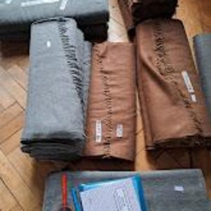 May include: Stacks of folded blankets in various shades of gray and brown. Some blankets have fringed edges. A red tape measure and a notepad with a blue cover are visible in the foreground, suggesting a textile or fabric product.