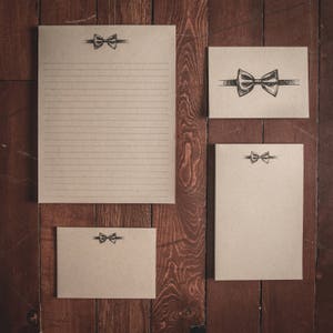 Pode incluir: Conjunto de três cartões de papelaria em branco com um design de gravata borboleta. Os cartões são feitos de papel castanho e têm uma área de escrita com linhas.