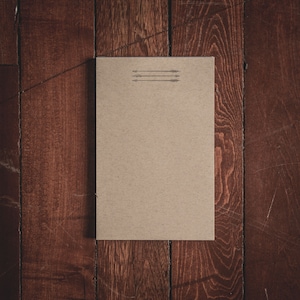 Könnte beinhalten: Ein leeres, braunes Papiernotizbuch mit drei kleinen Pfeilen, die obenauf gedruckt sind.