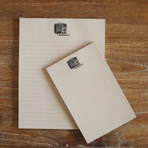 May include: Two blank lined notepads with a vintage camera graphic on each. The notepads are on a wooden surface.