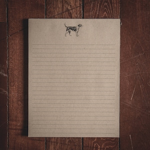 May include: A blank, lined notepad with a black and white illustration of a dog on the top. The notepad is on a wooden surface.