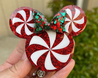 May include: Red and white peppermint candy cane themed Minnie Mouse ears with a green glitter bow. The ears are attached to a clear plastic badge holder.