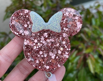 May include: A rose gold glitter Minnie Mouse badge holder with a white glitter bow. The holder is attached to a clear retractable badge reel.