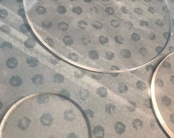 Peut inclure: Gros plan sur une surface grise avec un motif répétitif de petits cercles noirs. Trois sous-verres ronds en verre transparent sont placés sur la surface.