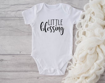 Peut inclure: Combinaison blanche pour bébé avec un texte noir qui dit "LITTLE blessing". La combinaison est posée sur une surface en bois clair avec une couverture en tricot blanc à droite.
