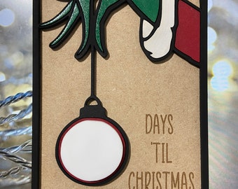 Peut inclure: Une main verte avec un poignet rouge tient un ornement de Noël rouge et blanc avec un cercle blanc au centre. Le texte "DAYS 'TIL CHRISTMAS" est sous l'ornement.