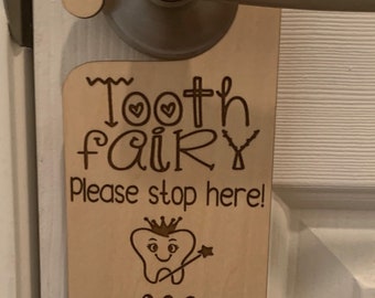 Peut inclure: Un porte-manteau en bois pour la fée des dents avec le texte "Tooth fairy please stop here!" et une dent de dessin animé avec une couronne et une baguette magique. Le nom "Mya" est écrit sous la dent avec le texte "Lost a tooth!"
