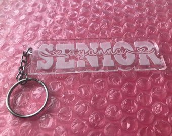 Peut inclure: Un porte-clés en acrylique transparent avec le mot "SENIOR" en lettres blanches et le nom "ayanna" écrit en cursive avec un cœur au-dessus.