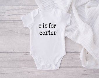 Peut inclure: Combinaison bébé blanche avec un texte noir qui dit "C est pour Carter".