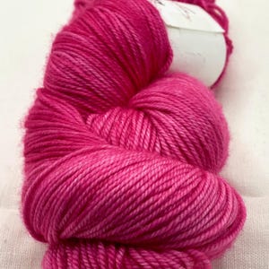 May include: A close-up shot of a vibrant fuchsia yarn skein. The yarn is tightly wound, revealing individual strands. A white label is partially visible. The yarn is a solid color, with subtle variations in tone, suggesting a high-quality fiber for knitting or crochet projects.