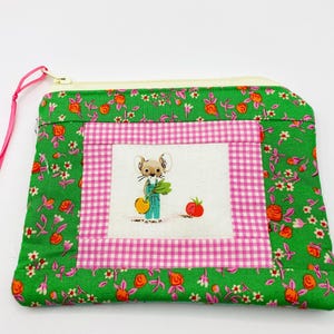 May include: A green floral fabric pouch with a pink zipper and a pink ribbon pull. The pouch features a central square with a mouse illustration holding vegetables, framed by pink gingham and green floral fabric.