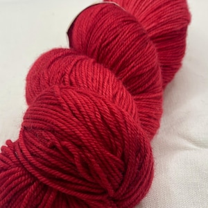 May include: A skein of vibrant red yarn, tightly wound and ready for knitting or crochet projects. The yarn is a solid, rich red color, with visible texture from the individual strands. The yarn is displayed on a white surface.