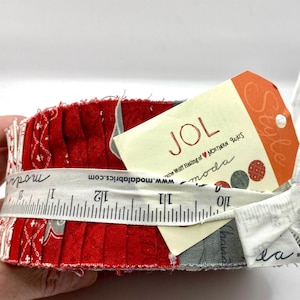 May include: A bundle of red fabric squares with a white measuring tape wrapped around it. A tag with the word "JOL" and the text "Style" is attached. The measuring tape shows inches.