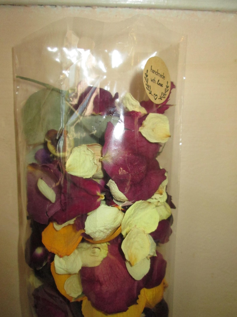 Puede incluir: Una bolsa transparente llena de p&eacute;talos de rosa secos en tonos burdeos, crema y amarillo. Una peque&ntilde;a pegatina ovalada con las palabras "handmade with love" est&aacute; adherida a la bolsa. Los p&eacute;talos parecen ser una mezcla de diferentes tipos de rosas.