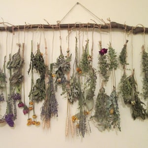 May include: A wall hanging made of dried flowers and herbs, with a rustic wooden branch at the top and twine strings holding the botanicals.