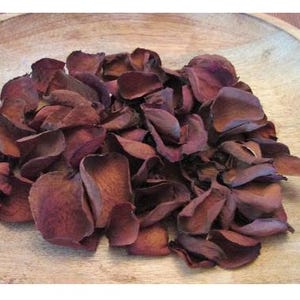 Puede incluir: Un mont&oacute;n de p&eacute;talos de rosa secos, de color burdeos, en un cuenco de madera marr&oacute;n claro. Los p&eacute;talos muestran una superficie texturizada con diversos tonos burdeos y marr&oacute;n. Los p&eacute;talos est&aacute;n dispuestos de forma informal, dando un aspecto natural y org&aacute;nico.