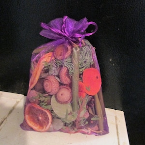 May include: A purple organza drawstring bag filled with dried potpourri, including brown acorns, green pine needles, orange citrus slices, and cinnamon sticks. The sheer fabric allows the contents to be visible through the mesh material.