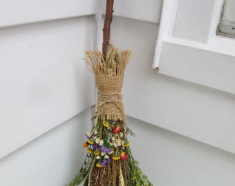 May include: A decorative broom constructed from a brown wooden branch handle and a bundle of dried flowers and grasses. The bristles are secured with a tan hessian wrap and tied with a matching twine bow, featuring yellow, purple, and red blooms.