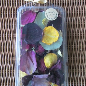 Puede incluir: Un recipiente de pl&aacute;stico transparente lleno de p&eacute;talos de flores secas de colores. Los p&eacute;talos son de varios tonos de morado, amarillo y verde. Una peque&ntilde;a pegatina redonda en el recipiente dice "handmade with love". El recipiente est&aacute; atado con una cinta dorada.