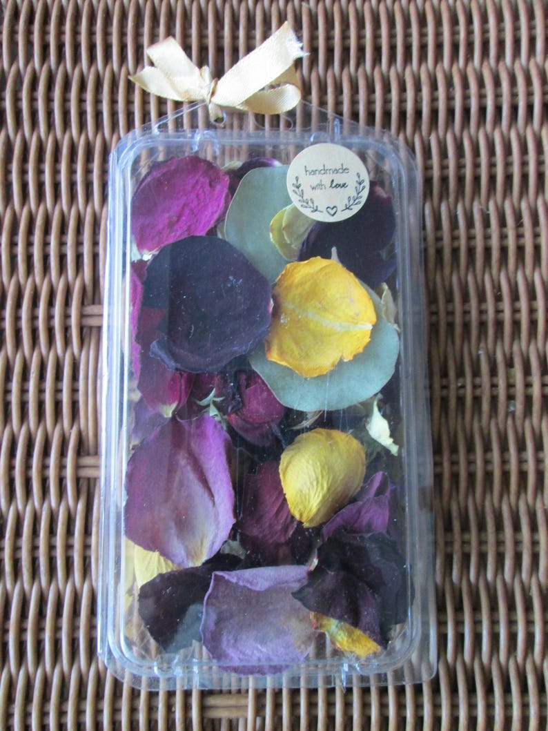Puede incluir: Un recipiente de pl&aacute;stico transparente lleno de p&eacute;talos de flores secas de colores. Los p&eacute;talos son de varios tonos de morado, amarillo y verde. Una peque&ntilde;a pegatina redonda en el recipiente dice "handmade with love". El recipiente est&aacute; atado con una cinta dorada.