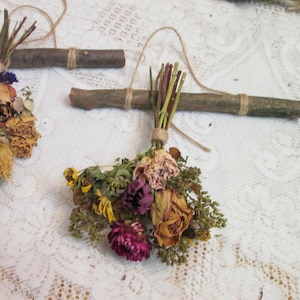 May include: A wall hanging decoration consisting of a small bundle of dried flowers in shades of yellow, pink, and deep purple, tied with twine to a natural brown wooden twig. The arrangement features a loop of twine for hanging purposes.