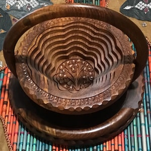 Op de afbeelding: Een houten fruitschaal met een gesneden ontwerp en een handvat. De schaal is gemaakt van donkerbruin hout en heeft een ronde vorm. De schaal staat op een kleurrijke mat.
