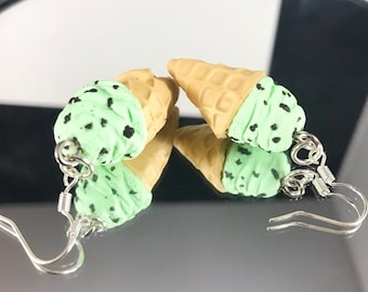 May include: A pair of green mint chip ice cream cone earrings with black chocolate chips. The earrings are made of polymer clay and have silver hooks.
